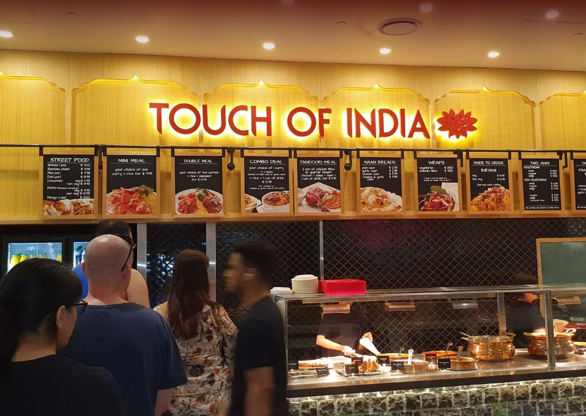 Customers queuing at a Touch of India food court counter under the illuminated menu boards